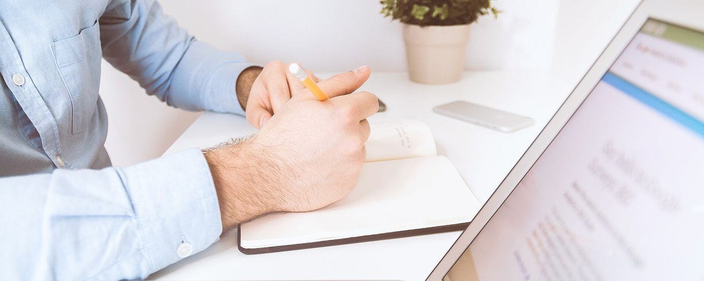 A close-up of a person writing in a notebook beside a laptop at a desk