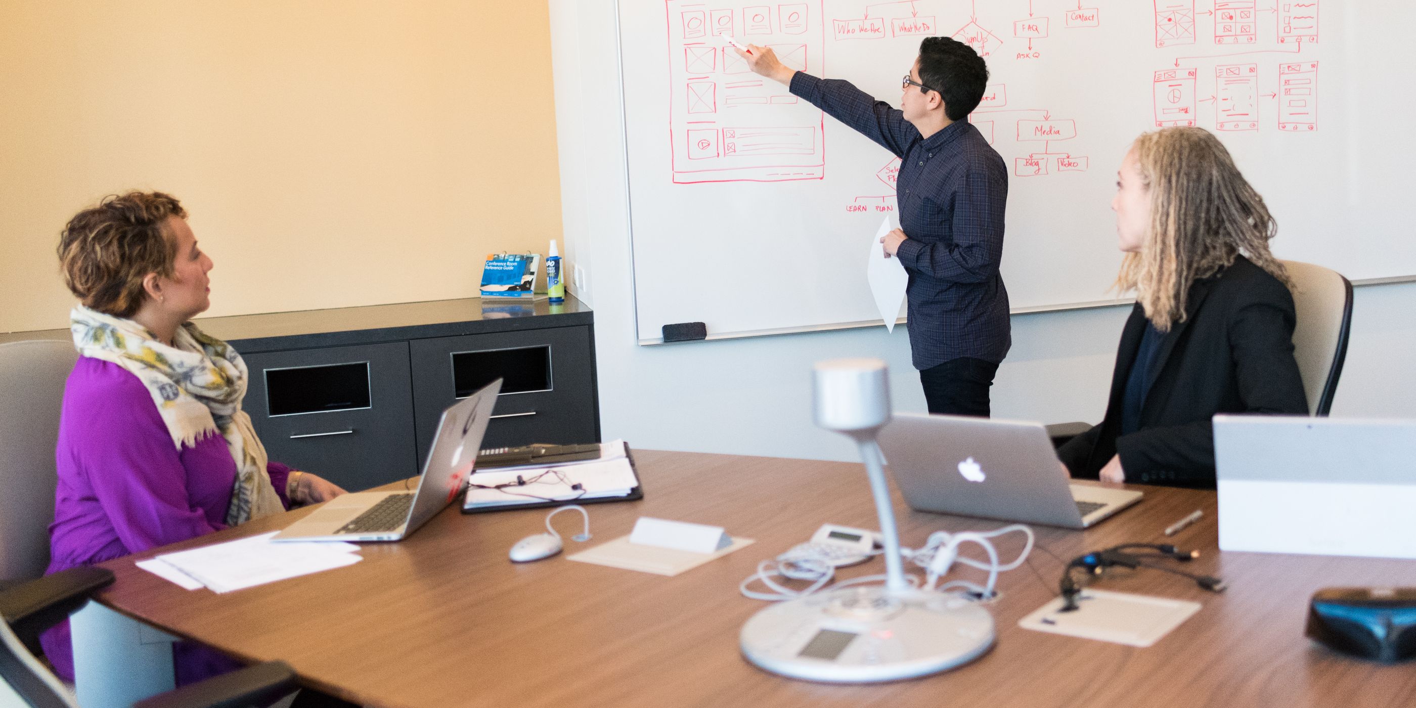A woman presenting a diagram on a whiteboard to two colleagues at a table