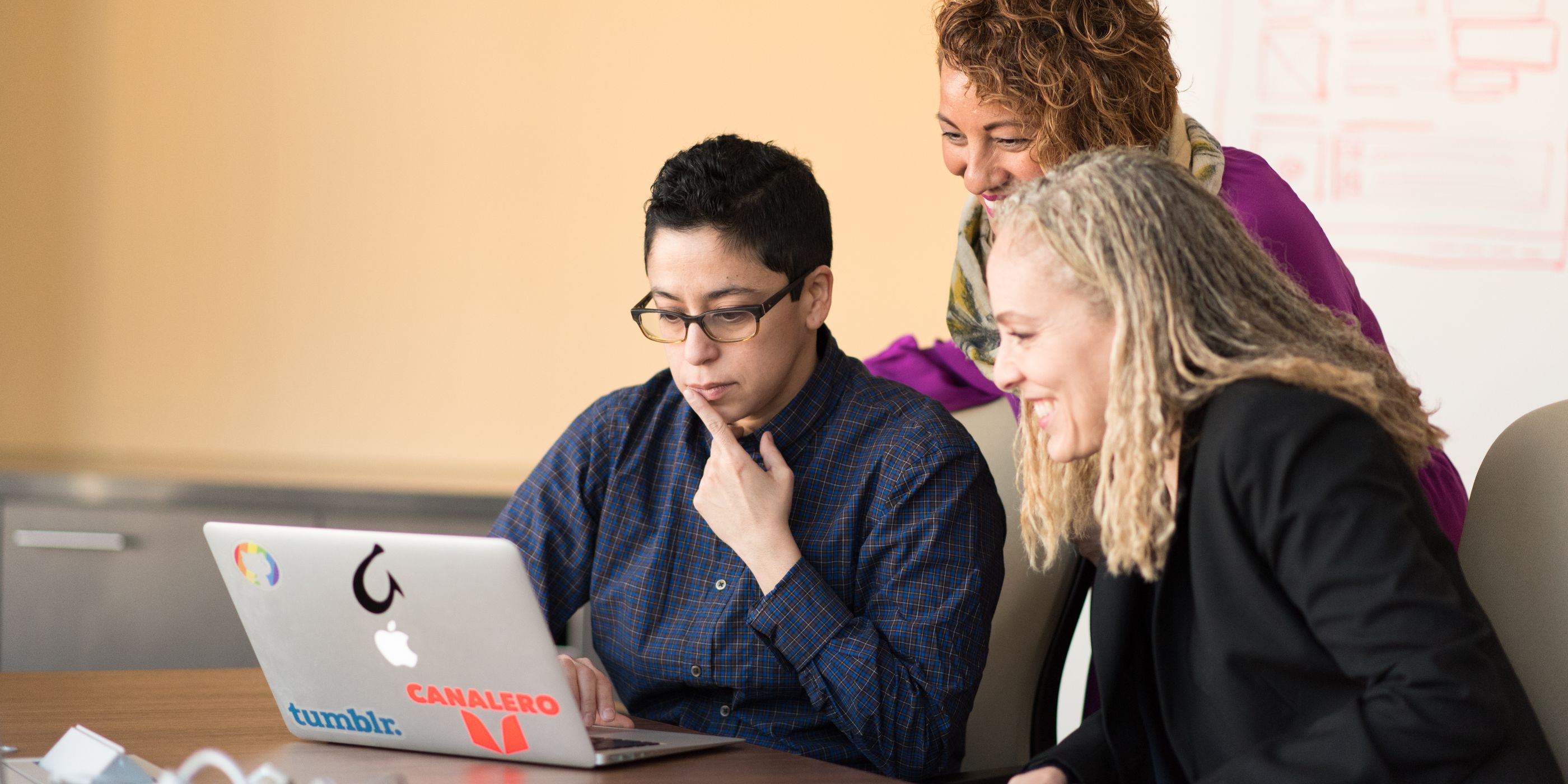 Three colleagues gathered around one laptop, a whiteboard behind them